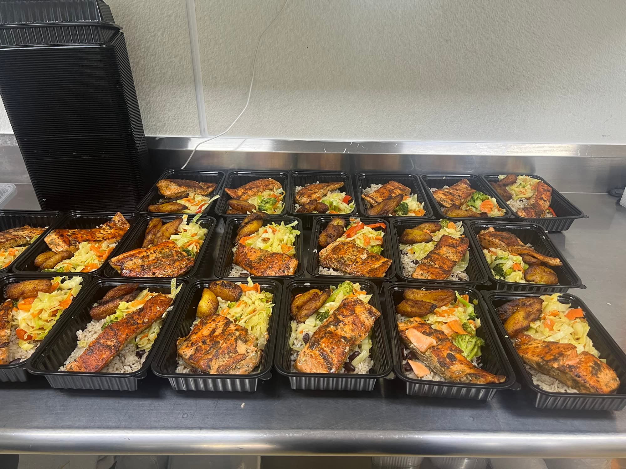 Rows of packed meal-prep containers with grilled salmon, rice and peas, cabbage slaw, and fried plantain